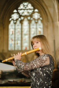 Annabel Knight playing flute, standing in front of decorated harpsichord lid, with stained glass windows in the background
